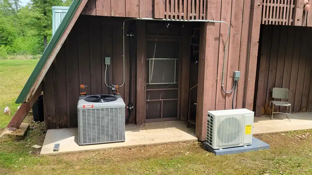 A new condenser and a ductless mini-split outdoor unit installed on a pad beside a dark-stained wooden A-frame building