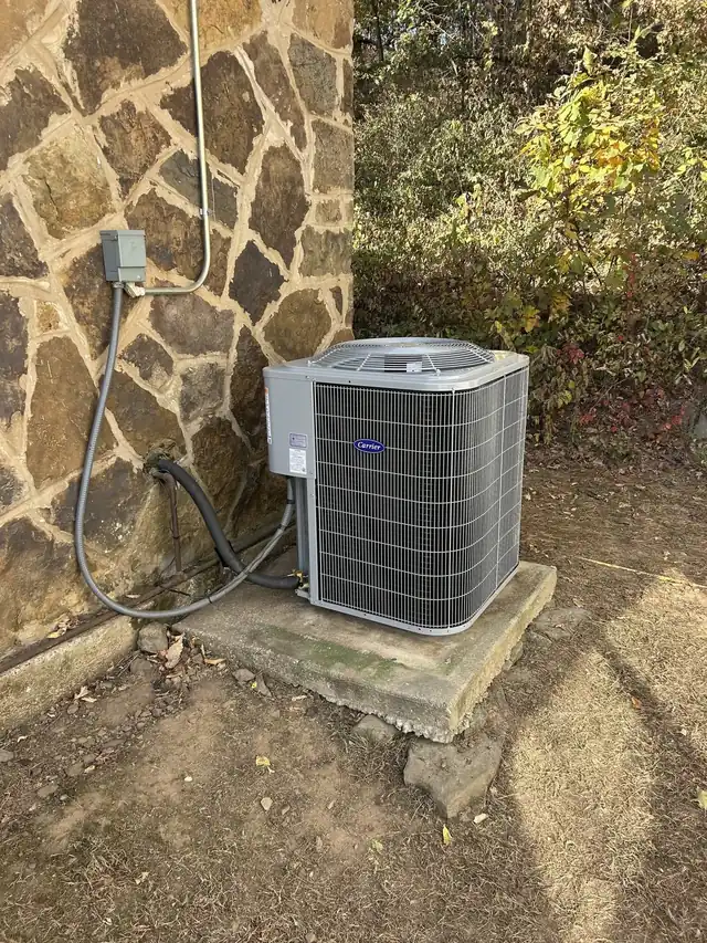 A new Carrier condenser on a concrete pad against a fieldstone wall with autumn woods behind