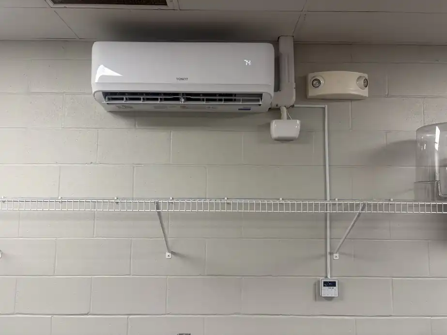 A ductless mini-split head mounted on a painted block wall above a wire shelf, with its line set and thermostat