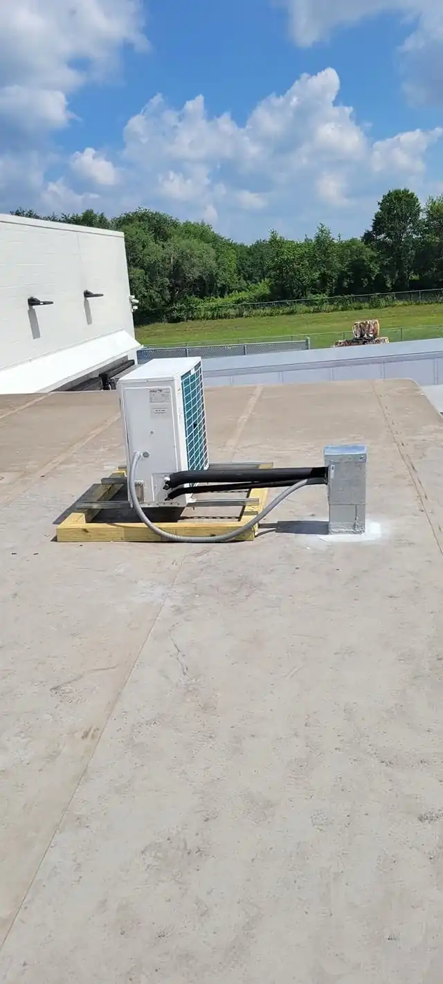A mini-split outdoor unit on a timber stand on a flat roof, photographed from the parapet side with the line set running along the roof
