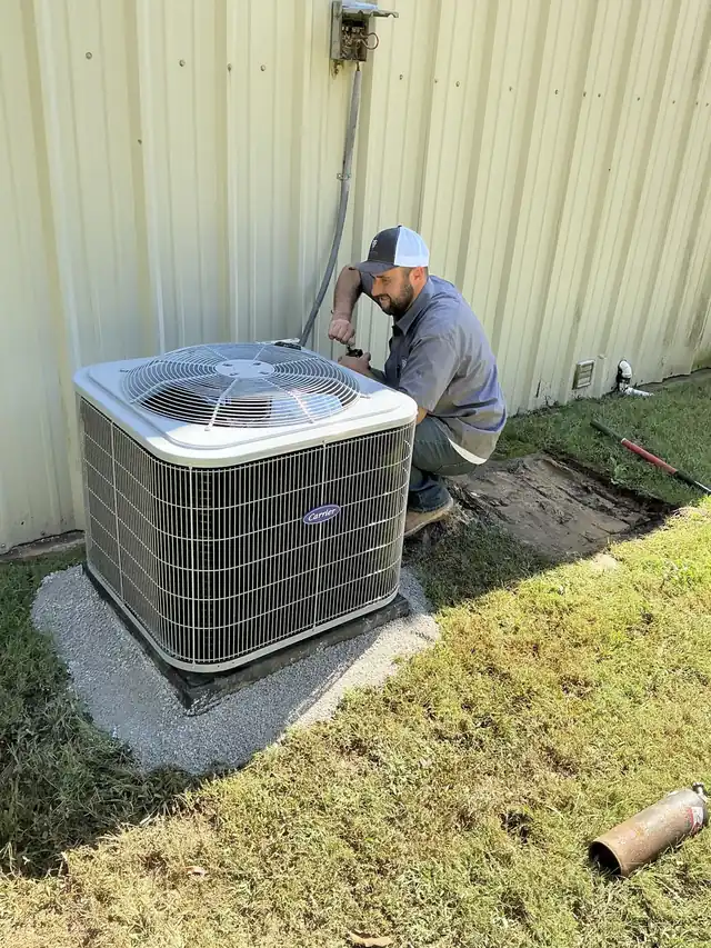 A technician in a Trinity shirt kneeling at a new Carrier condenser beside a metal building