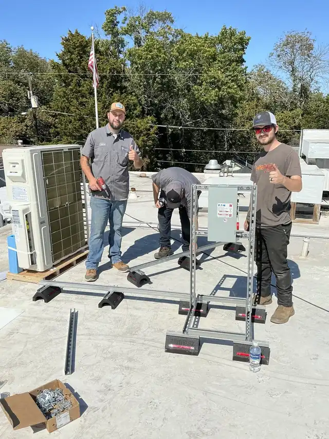 Three Trinity technicians on a commercial roof partway through a mini-split install on a strut stand, two giving a thumbs-up, a US flag behind them
