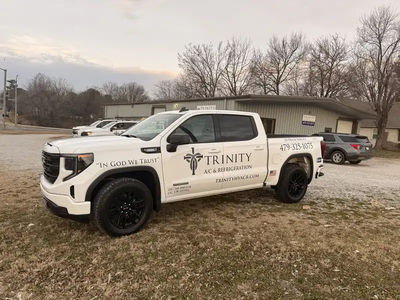 A white Trinity GMC pickup lettered In God We Trust, with two more fleet trucks and the Springdale shop behind it at dusk